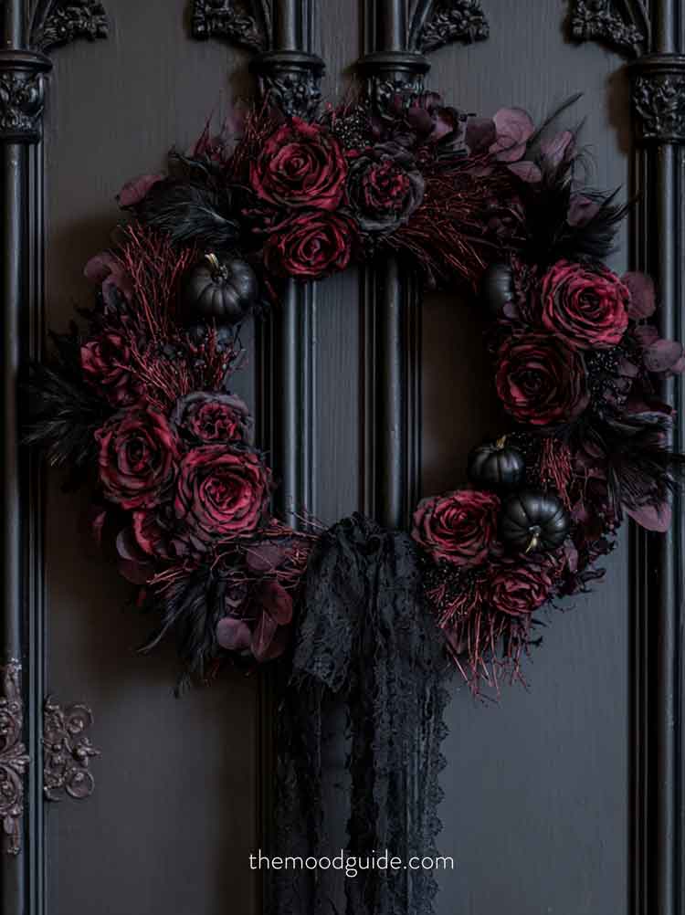 A moody, dramatic Dark Gothic fall wreath hanging on a matte black gothic wooden door with antique iron hardware. The wreath is composed of deep burgundy and black silk roses, dark charcoal-colored dried twigs, preserved black eucalyptus leaves, miniature matte black pumpkins, and dark feathers, wrapped in a tattered black lace ribbon. mysterious and enchanting 