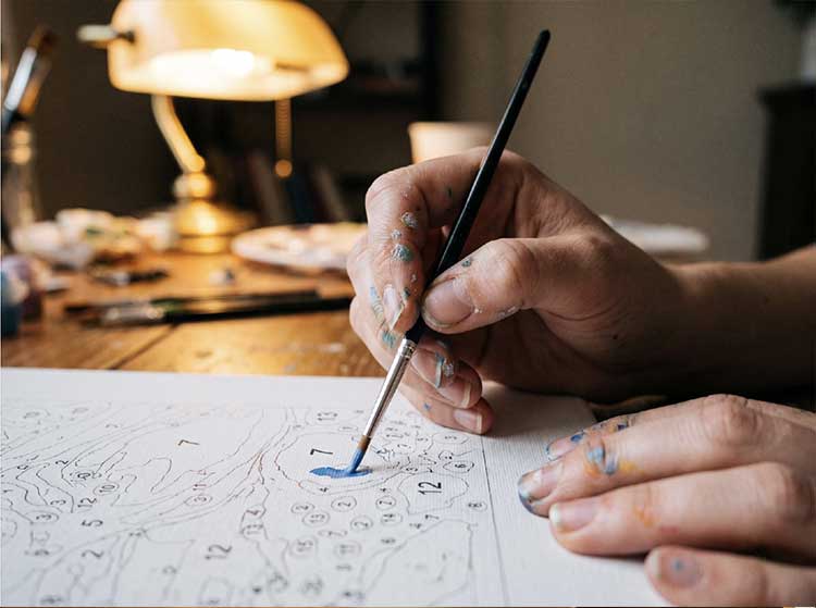 Close-up of hands dabbing paint onto a numbered canvas with a small brush, warm lamplight in the background
Hands work through a numbered canvas under warm lamplight.