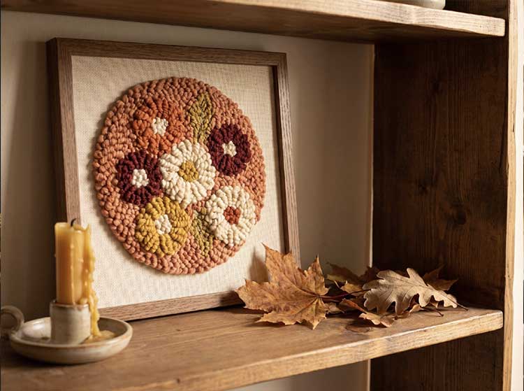 Framed punch needle floral piece with looped yarn texture resting on a shelf next to a candle and autumn leavesA looped-yarn floral piece adds texture to a fall shelf.
